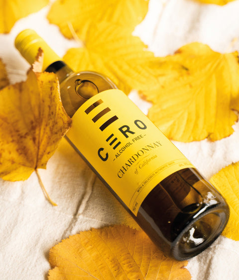 Een wijnfles 'CÈRO' alcoholvrije chardonnay liggend op een witte ondergrond met herfstbladeren.
