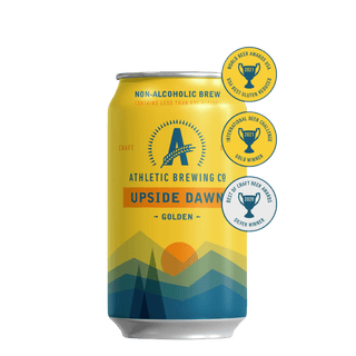 Een geel blikje Athletic Brewing's 'Upside Dawn' non-alcoholische Golden Ale met een design dat landschapselementen toont en omringd door gouden award-stickers.
