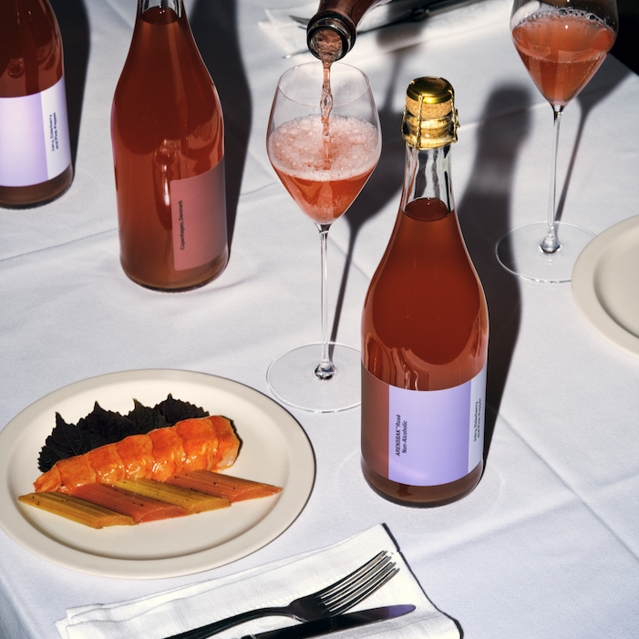 Fles alcoholvrije sparkling tea, roze drank in wijnglazen en kleurrijk gerecht op witte tafel | World of NIX
