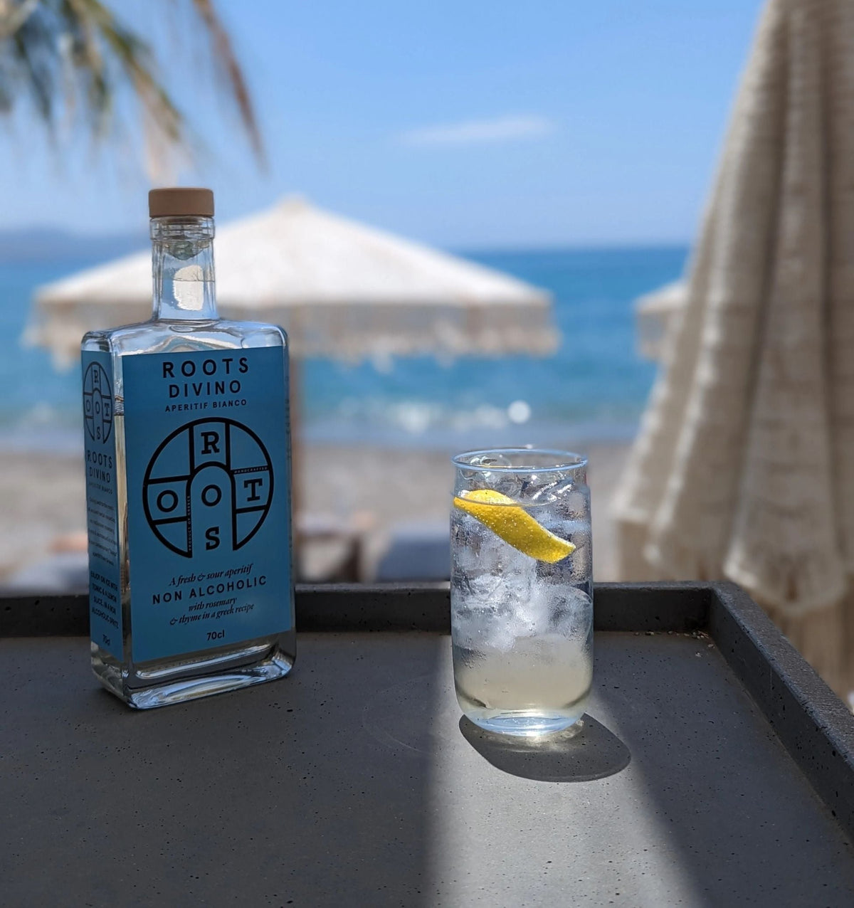 Fles Roots Divino Aperitif Bianco en alcoholvrije cocktail met citroen op strandtafel, zee op achtergrond | World of NIX