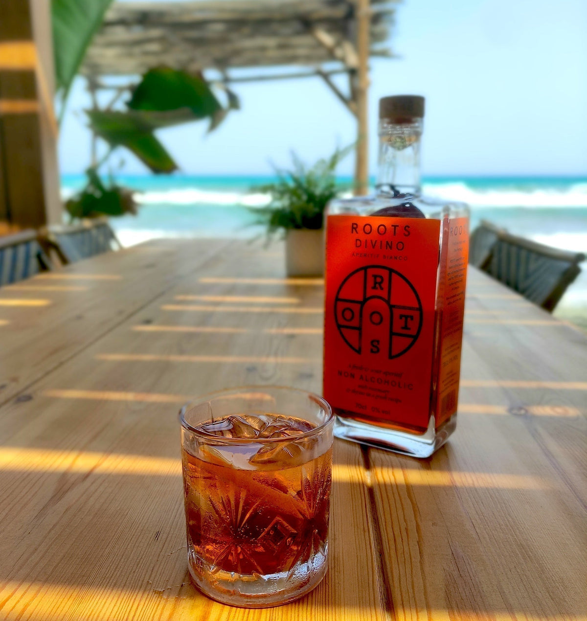Glas alcoholvrije Negroni met Roots Divino fles op houten tafel bij het strand | World of NIX
