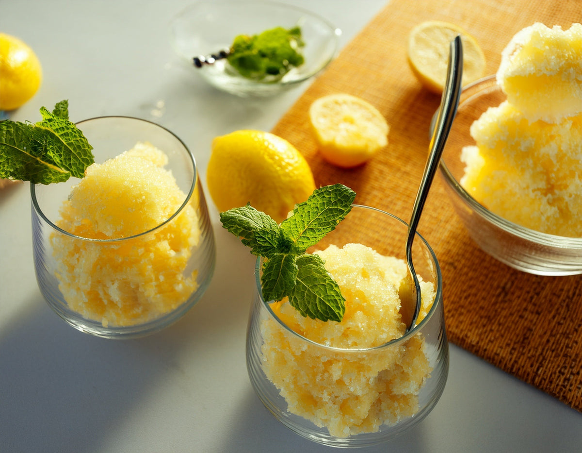 Twee glazen citroen granita met verse munt, citroenen en een kom granita op tafel | World of NIX
