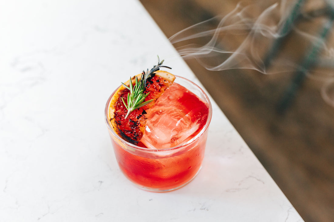 Alcoholvrije rode cocktail met grote ijsblok, gegrilde sinaasappel en rozemarijn op witte tafel | World of NIX