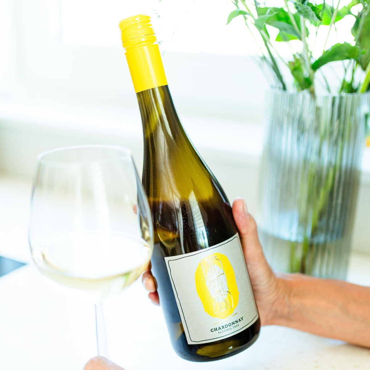 Fles alcoholvrije chardonnay met geel etiket naast wijnglas op tafel, bloemenvaas op achtergrond | World of NIX