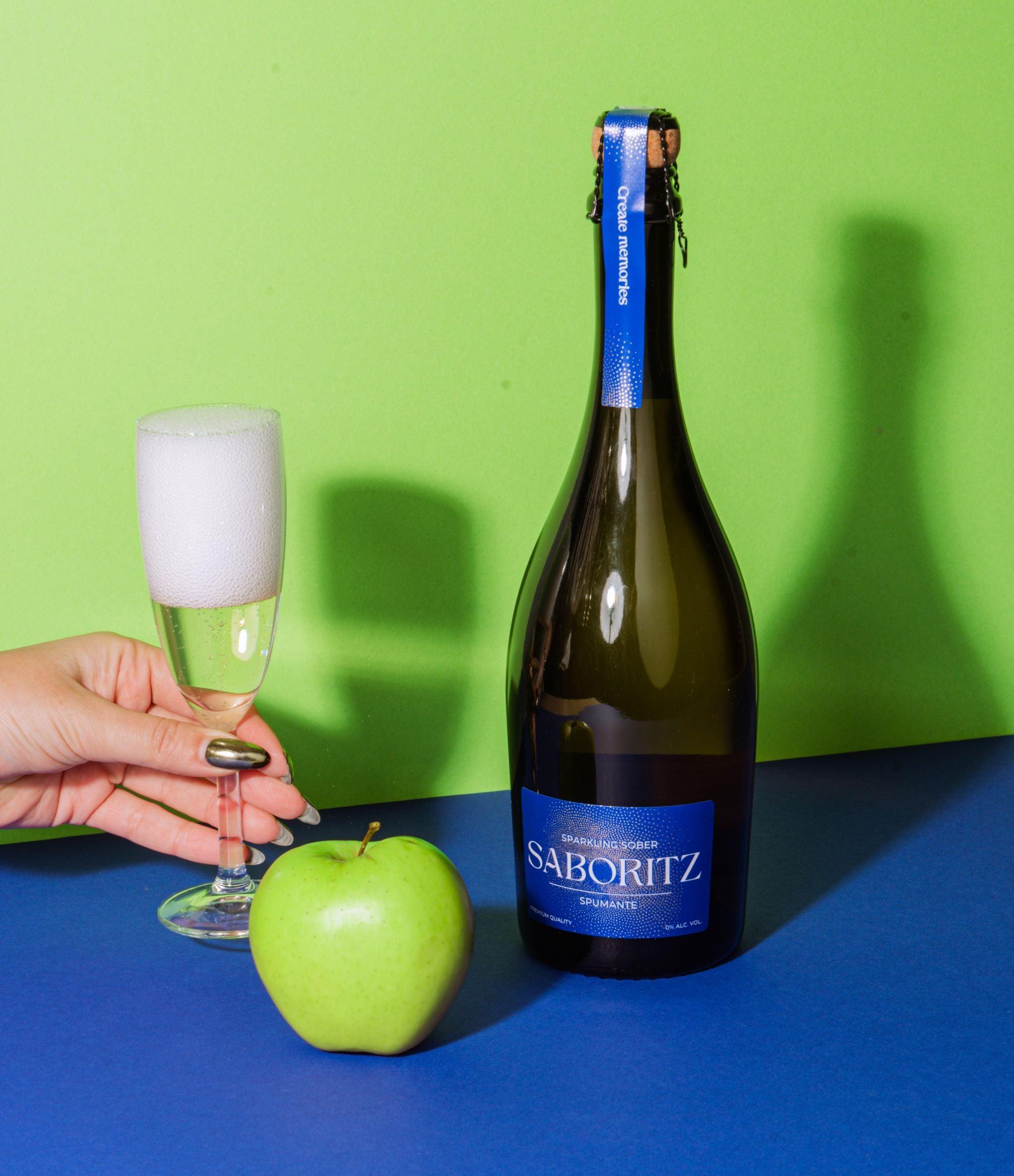 Fles alcoholvrije Saboritz spumante, glas met schuimend drankje en groene appel op blauwe ondergrond | World of NIX