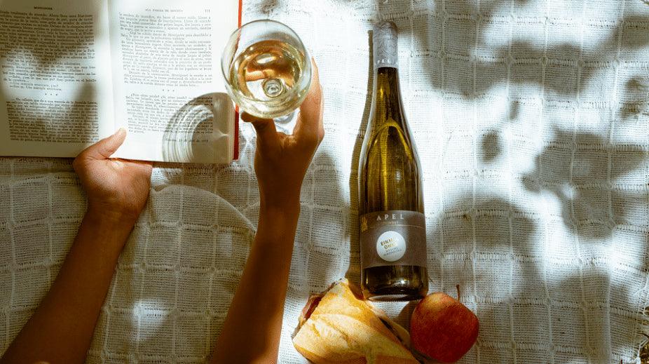 Boek lezen met glas alcoholvrije wijn, fles Apel, stokbrood en appel op kleed | World of NIX