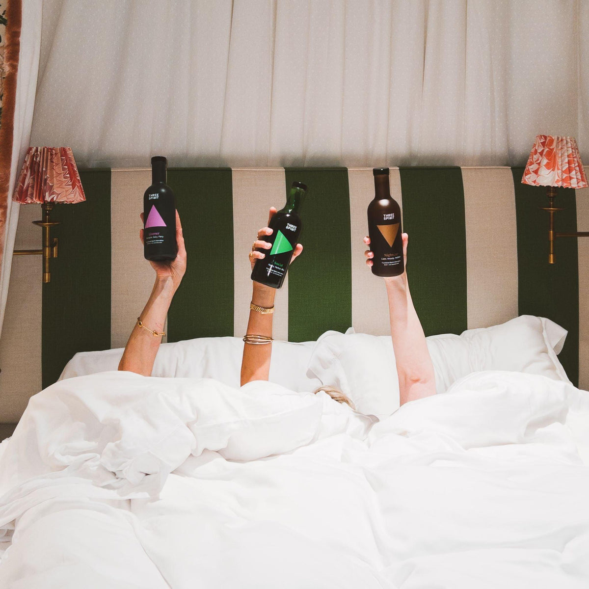 Drie handen in bed houden Three Spirit alcoholvrije spirits omhoog, groene gestreepte hoofdbord | World of NIX