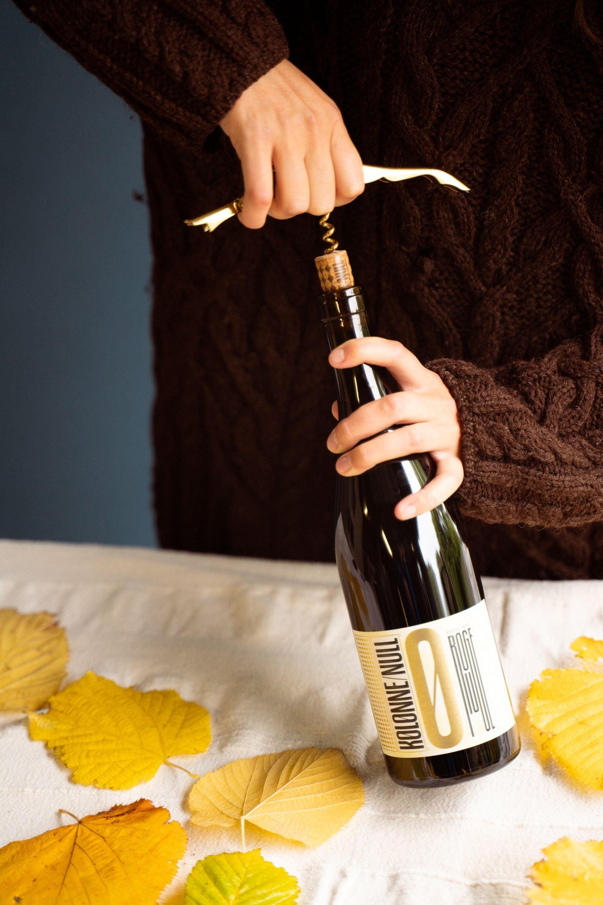 Hand opent fles alcoholvrije wijn met kurkentrekker, herfstbladeren op tafel | World of NIX