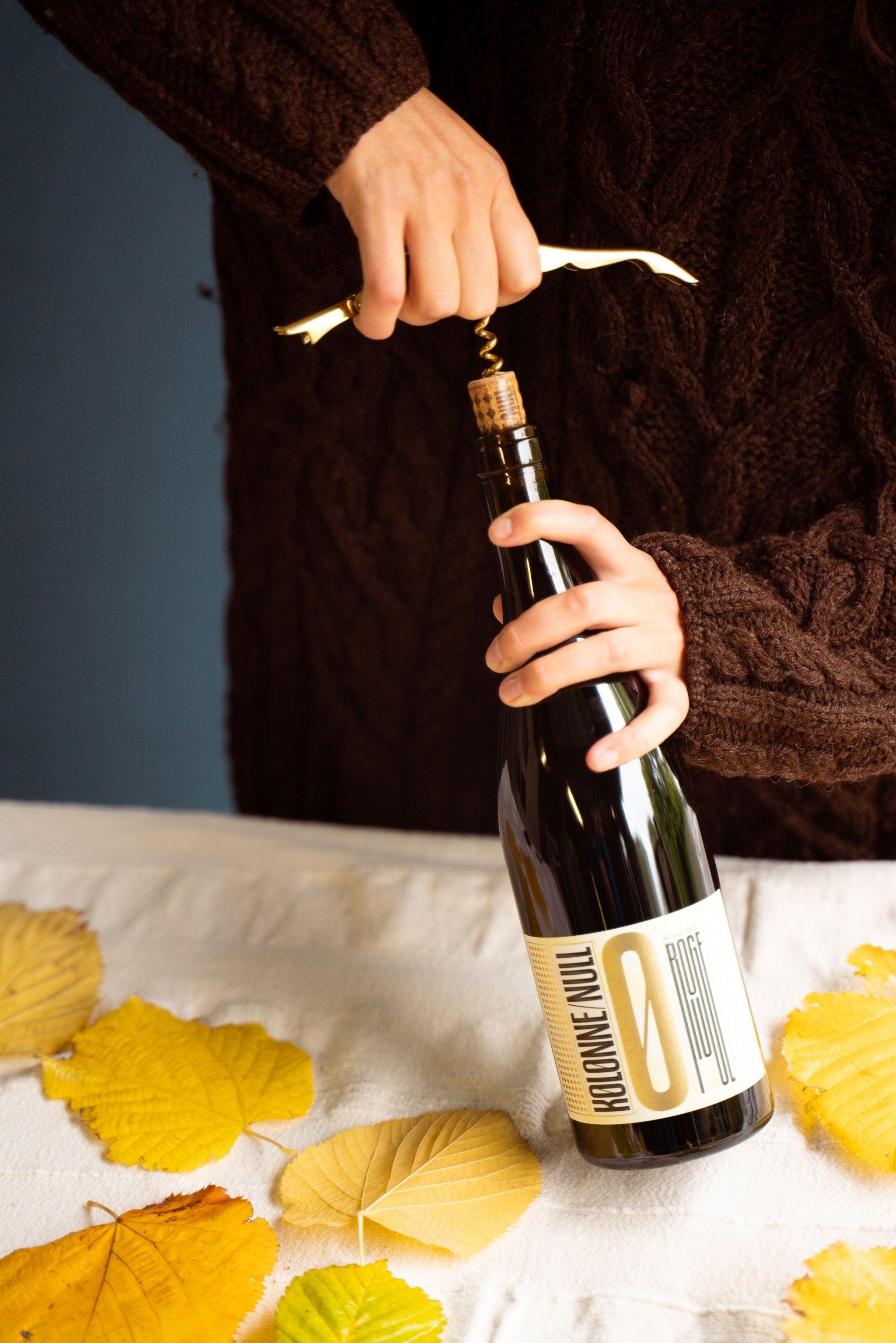Hand opent fles alcoholvrije wijn met kurkentrekker, herfstbladeren op tafel | World of NIX