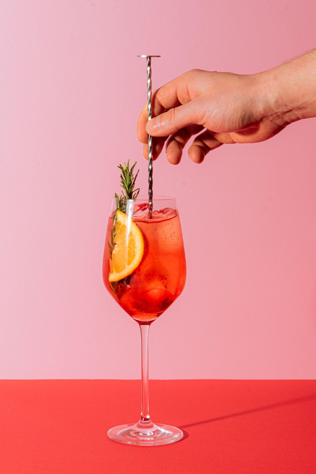 Hand roert alcoholvrije cocktail met sinaasappel en rozemarijn in wijnglas op roze achtergrond | World of NIX