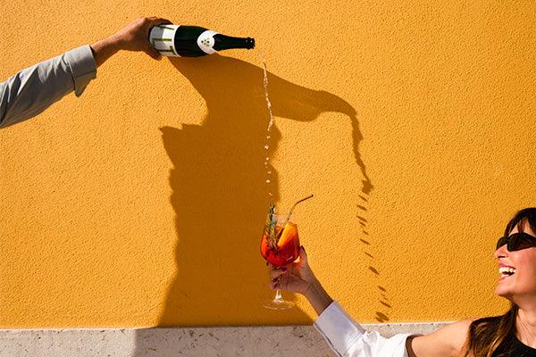 Vrouw ontvangt sprankelende drank van fles in glas bij zonnige gele muur, zomer sfeer | World of NIX