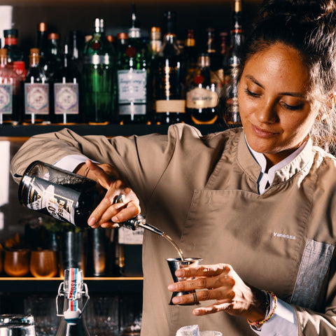 Een barvrouw, geconcentreerd op het schenken in een jigger uit een fles REBELS 0.0% Malt Blend, met op de achtergrond een bar vol flessen.