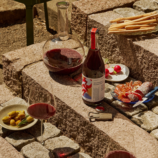 Een picknick setting met een fles Ambijus cider en twee glazen gevuld met de Ambijus, gepresenteerd op een stenen trap naast een bord met olijven en fuet.