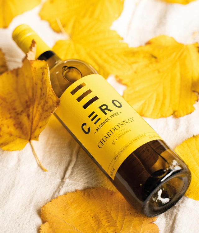 Een wijnfles 'CÈRO' alcoholvrije chardonnay liggend op een witte ondergrond met herfstbladeren.