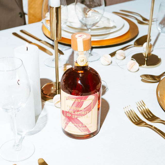 Een elegante eettafel met witte servetten, zilver bestek, en een fles Rebels 0.0% Sweet Amaretti midden op de tafel tussen de glazen.