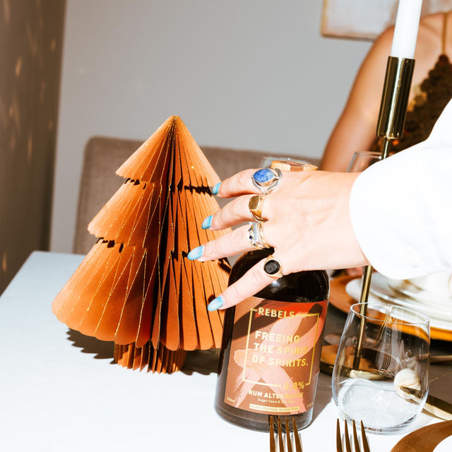 Een feestelijk gedekte tafel met een fles Rebels 0.0% Dark Spice, gevouwen servetten en gouden bestek, klaar voor een evenement of viering.