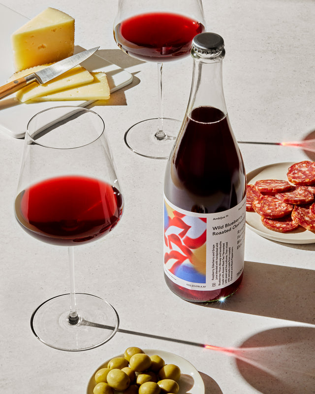 Een geopende fles Ambijus cider met een donkerrode tint naast twee elegante wijn glazen op een witte ondergrond, vergezeld van een bord met olijven en stukken kaas.