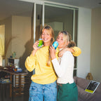 Twee personen, lachend en proostend met glazen bier, in een huiselijke setting met een onscherpe achtergrond.