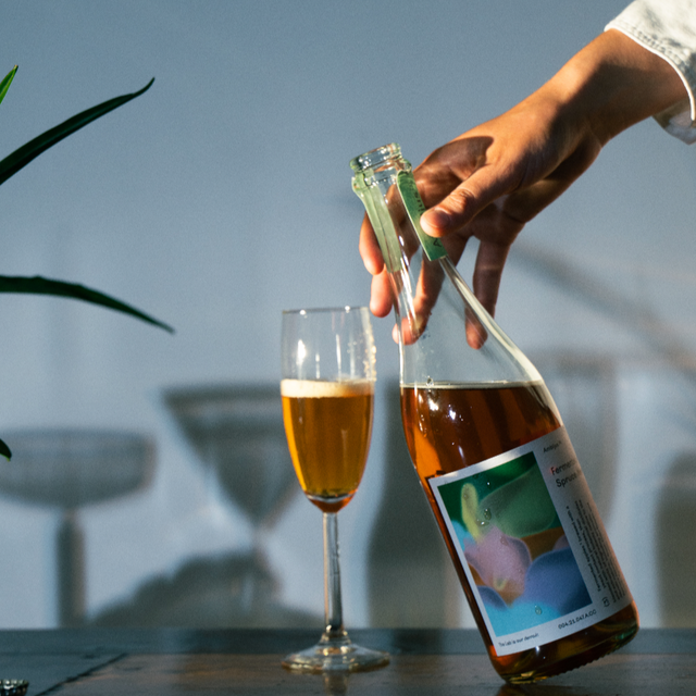Een glas wordt gevuld met Ambijus cider, helder en sprankelend, met een persoon die de fles vasthoudt tegen een vage achtergrond van een buitenomgeving.