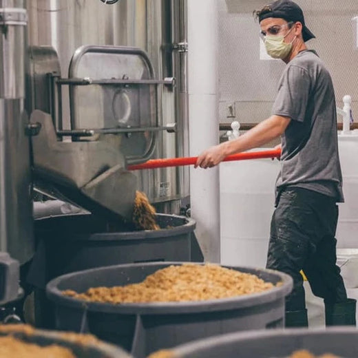 Een brouwer in een tanktop werkt aan het brouwproces in een brouwerij, met een focus op een grote roestvrijstalen brouwketel.